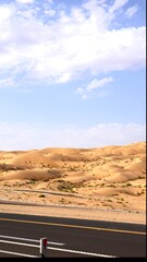 Curving asphalt road threads across wind-brushed dunes under painted sky, bright yellow line holding its course through soft sand that creeps onto shoulder, showing tension between nature and