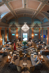 Elegant lounge featuring a grand chandelier, plush seating, checkered floor, and a woman in a red dress walking through the sophisticated space.