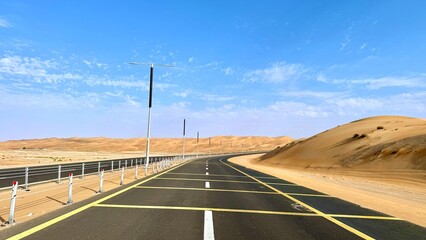Road slices through golden sand as poles line both sides, road reflects heat under vibrant sky while sand dunes rise gently, creating harmony between built line and desert stillness.
