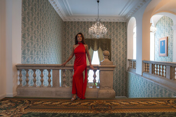 A woman in a red dress stands by a stone balustrade in a luxury hotel interior with a chandelier,...