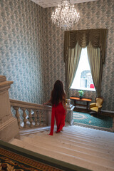 A beautiful woman in a red dress descends a marble staircase with a classic balustrade. Floral...