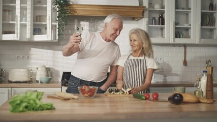 Old people and modern technology, aged man filming by smartphone in home kitchen . Charming elderly woman cooking dinner and her husband holding smartphone, video call or online streaming in internet - Powered by Adobe