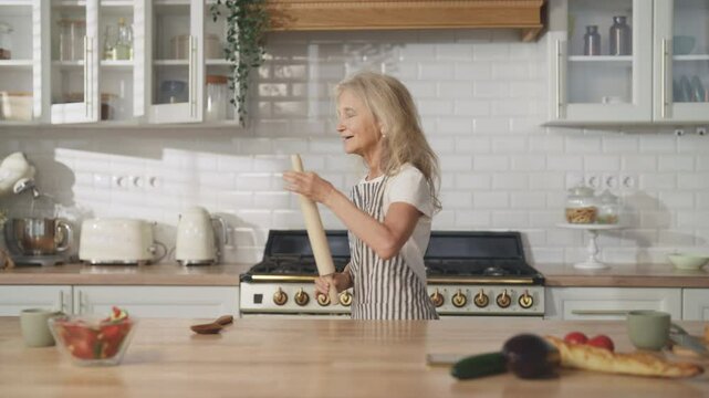 Carefree old woman enjoying music and dancing alone in home kitchen, portrait. Charming elderly lady singing into wooden rolling-pin ,good mood and positivity, anti-stress and recharge with energy