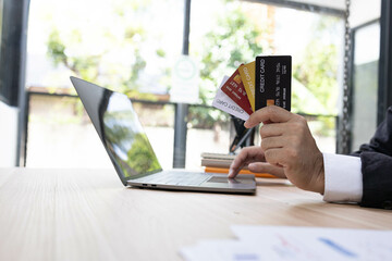 A man is using a laptop and a cell phone to pay for something. He is holding a credit card and a debit card
