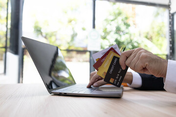 A man is using a laptop and a cell phone to pay for something. He is holding a credit card and a debit card
