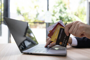 A man is using a laptop and a cell phone to pay for something. He is holding a credit card and a debit card
