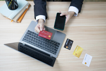 A man is using a laptop and a cell phone to pay for something. He is holding a credit card and a debit card

