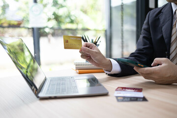 A man is using a laptop and a cell phone to pay for something. He is holding a credit card and a debit card
