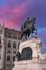 Obraz premium An equestrian statue of Ferenc Rakoczi II on an ornate pedestal stands before the neo Gothic Hungarian Parliament Building, with a pink and purple sky in the background.