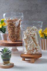 Solondok is traditional Indonesian crispy snack, displayed on a small wooden pedestal. Another similar pouch with a different snack is fried Makaroni, seen in the background. The setup creates a clean