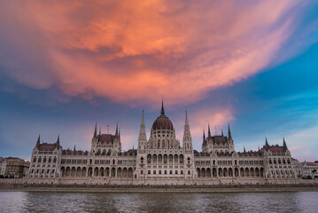Fototapeta premium The neo Gothic Hungarian Parliament Building in Budapest, Hungary, stands by the Danube River under a vibrant orange and pink sky with calm reflections.