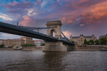 Fototapeta premium The Szechenyi Chain Bridge spans the Danube River in Budapest, Hungary, with stone towers, suspension cables, and a vibrant sunset sky reflected in the water.