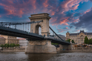 Obraz premium The Szechenyi Chain Bridge spans the Danube River in Budapest under a colorful sunset, with stone towers, suspension cables, and a historic building visible.