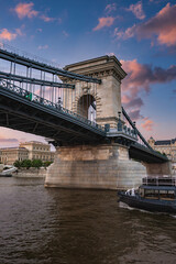 The Szechenyi Chain Bridge spans the Danube River in Budapest, Hungary, with stone towers, suspension cables, and a vibrant sunset sky. A boat is on the river.