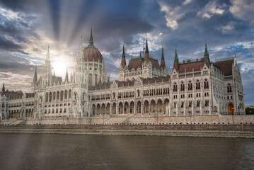 Obraz premium The neo Gothic Hungarian Parliament Building stands by the Danube River in Budapest, with sunlight streaming through clouds and calm waters in the foreground.