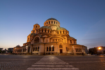 Fototapeta premium Alexander Nevsky Cathedral in Sofia illuminated by warm sunset light, emphasizing its grand Neo-Byzantine architecture and majestic domes under a clear evening sky with a quiet street view.