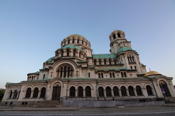 Fototapeta premium Wide frontal view of Alexander Nevsky Cathedral in Sofia at dusk, highlighting its grand Neo-Byzantine architecture, green domes, and intricate stone facade under a clear evening sky.