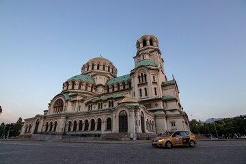 Fototapeta premium Alexander Nevsky Cathedral in Sofia at dusk with a yellow taxi in the foreground, highlighting its grand Neo-Byzantine architecture, green domes, and intricate stone facade under a clear sky.