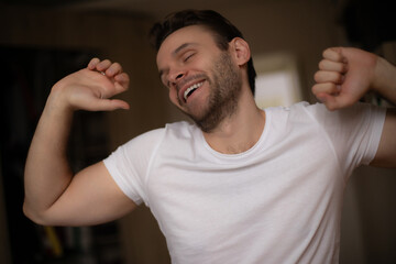 Happy young man in a white T-shirt stretching and smiling with eyes closed, enjoying a fresh...