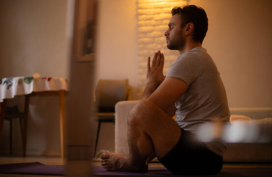 Man practicing yoga at home in a seated meditation pose, hands in prayer position, wearing a t-shirt and shorts, in a peaceful living room with warm lighting and calm atmosphere.