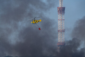 A yellow firefighting helicopter drops water over heavy smoke after a missile strike in Kyiv, with a TV tower in the background during the Russia-Ukraine war.