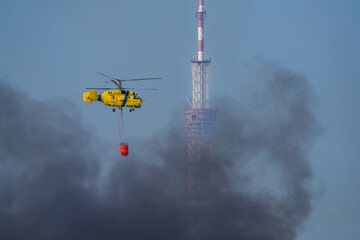 A yellow firefighting helicopter drops water over heavy smoke after a missile strike in Kyiv, with a TV tower in the background during the Russia-Ukraine war.