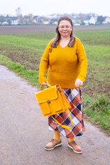 Stylish mature woman in cozy yellow sweater with handbag standing on rural road, elegance retro style, and nature. Countryside life and autumn fashion, yellow bag, outdoor lifestyle