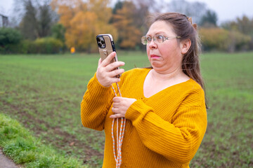 Mature woman scrolling smartphone, surprised by online news, sitting indoors, wasting time, casual clothing, emotional face