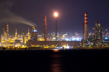 Night view of an illuminated oil refinery with flaming smokestack and smoke emissions, industrial chimneys and lights reflecting energy production and environmental concerns by the coastline.