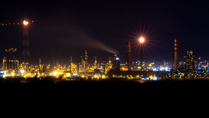 Fototapeta premium Night view of an illuminated oil refinery with glowing chimneys, industrial buildings, and flaring towers under a dark sky, showcasing the energy and manufacturing sector's nighttime operations.