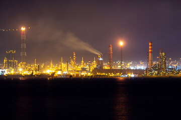 Naklejka premium Brightly lit oil refinery at night with smoke emissions, flaring towers, and glowing chimneys against a dark sky, illustrating industrial activity and energy production in full operation.