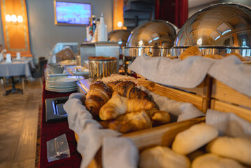 A breakfast buffet with croissants, bread rolls, and silver chafing dishes in a cozy dining area with ornate sconces and neatly arranged tables.