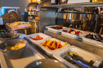 A high end buffet features fresh vegetables, stuffed peppers, and olives in white trays, with metallic chafing dishes and warm lighting in the background.