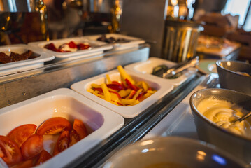 A buffet setup in a luxury boutique hotel features sliced tomatoes, bell peppers, condiments, and sauces in white trays under warm lighting.