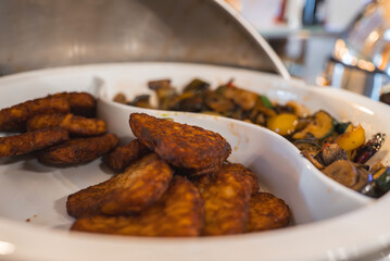 A white serving dish with golden hash browns and colorful sauteed vegetables, part of a buffet setup in warm lighting with a metallic lid in view.