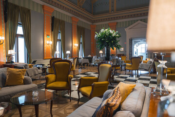 Spacious lounge with high ceilings, tall windows, green and gold drapes, orange columns, checkered marble floor, plush seating, and floral centerpiece.