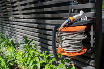 Garden hose with nozzle neatly wound on reel attached to wooden wall of house terrace on sunny day