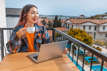 Young woman working remotely on vacation, enjoying a cup of coffee on a balcony with a beautiful sea view
