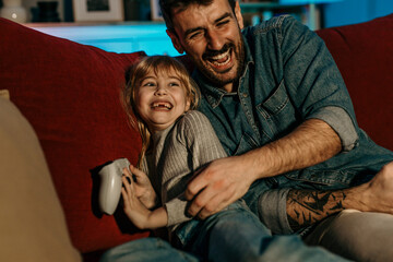 Obraz premium Father and daughter laughing and playing video games on sofa at home