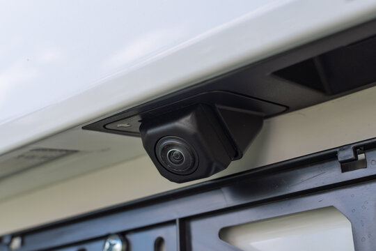 A modern backup camera is mounted near license plate, enhancing driving safety and parking precision on a white car.