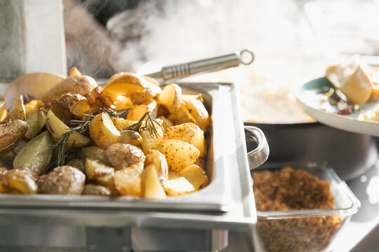 Close-up of roasted potatoes with rosemary in a buffet tray, with steam rising in a warm catering setting.