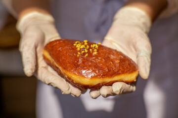 Hand in glove holding a traditional Polish donut with orange glaze and crushed pistachios.