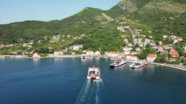 Aerial view of a passenger ferry sailing in the Bay of Kotor against the backdrop of a picturesque mountain coastline and a seaside town on a sunny day
