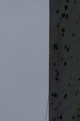 A textured climbing wall with colorful holds and a safety rope extends upward under a muted sky, conveying challenge and vertical adventure