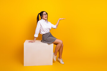 Young woman in casual uniform sitting on a box over yellow background making a welcoming hand...