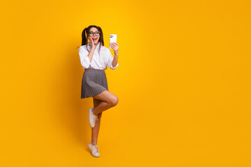 Smiling young woman holding a phone while posing against a vibrant yellow background, dressed...