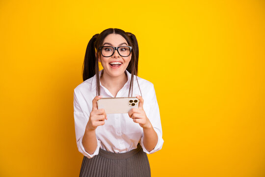Cheerful schoolgirl with twin ponytails using smartphone on vibrant yellow background in casual formal attire