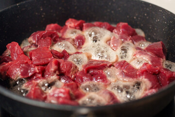 Chunks of raw beef are sizzling in a dark pan, with steam and bubbles rising from the meat as it cooks