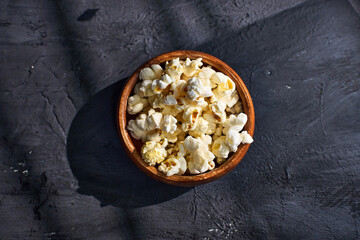 A wooden bowl filled with fluffy, freshly popped popcorn rests on a dark textured surface