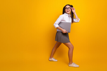 Young student with laptop walking confidently in stylish attire and glasses on vibrant yellow...